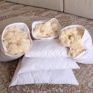 May include: A collection of white pillows featuring zipped openings that reveal a fluffy, cream-coloured wool or fibre fill. Two pillows are stacked flat, while three others are positioned upright with their zips partially open.