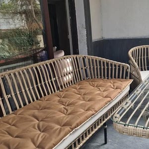 May include: A long, curved rattan sofa featuring a light natural wood finish and a grid-patterned frame. The seat is topped with a long, tufted, tan-colored cushion that rests on a thin white base pad, supported by slender black metal legs.