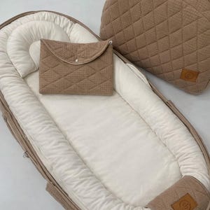 May include: A tan baby nest with a matching diaper bag and small envelope-style pouch. The nest has a soft, white interior and a quilted exterior. The diaper bag and pouch also feature a quilted design. The set is designed for baby care.