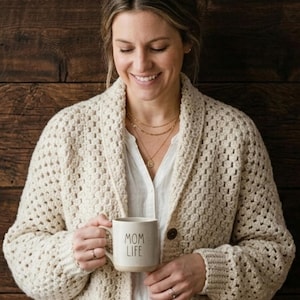 Puede incluir: Cárdigan de punto grueso color crema con grandes botones de madera y bolsillos. La mujer sostiene una taza que dice "MOM LIFE". Una cesta tejida con flores secas y algodón está a la izquierda. El fondo es una pared de madera oscura.