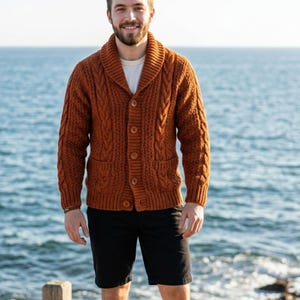 Può includere: Un cardigan in maglia a trecce color arancio bruciato con collo a scialle, maniche lunghe e chiusura frontale con bottoni e due tasche, indossato sopra una t-shirt bianca semplice e pantaloncini neri a tinta unita.