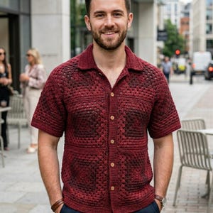 May include: A burgundy crochet shirt with short sleeves and a collared neckline. The shirt features a button-down front and two chest pockets. The open weave design reveals a darker underlayer, creating a textured appearance. The shirt is worn by a person.