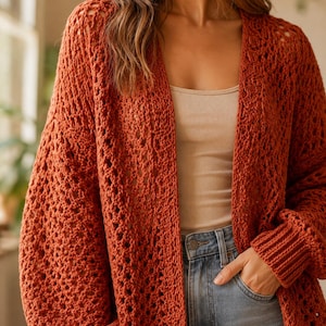 May include: A rust-coloured open-front cardigan featuring a loose, airy crochet knit pattern with long sleeves, ribbed cuffs, and a relaxed fit. The garment is worn over a simple beige tank top and light blue denim jeans with a visible metal button.
