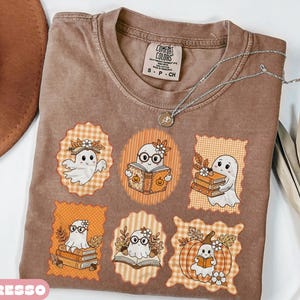 May include: A brown short-sleeved t-shirt featuring a Comfort Colors label and a graphic print of six orange-toned frames containing illustrations of ghosts reading books, wearing glasses, and sitting with pumpkins and autumn leaves.