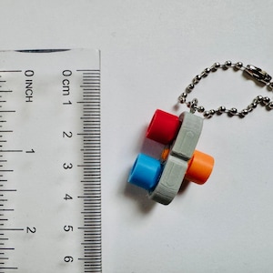 May include: A colorful keychain featuring a gray plastic base with red, blue, and orange cylindrical accents. The keychain is attached to a silver ball chain. The object is approximately 1 inch in length.