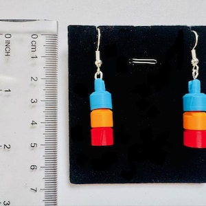 May include: A pair of colorful earrings with a stacked design. Each earring features a blue top, an orange middle section, and a red bottom. The earrings are suspended from silver-colored hooks. The ruler next to the earrings shows a measurement of approximately 1 inch.