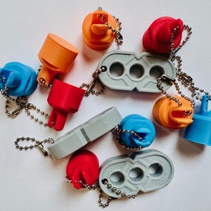 May include: A collection of colorful 3D-printed keychains. The keychains feature various shapes and designs, including cylindrical and geometric forms, in shades of blue, orange, and red. Some keychains have a gray, rectangular design with three holes.