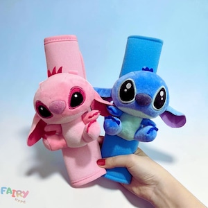 May include: Two plush seatbelt covers, one pink and one blue, featuring cartoon characters with large eyes and ears. The pink character has a crown and the blue character has a small crown. The seatbelt covers are held by a hand.