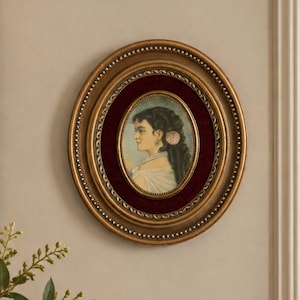 May include: An oval-shaped portrait painting of a person in profile, encased in a decorative gold frame with a deep red velvet mat. The artwork depicts a figure with dark hair and light clothing, mounted on a wall above a wooden surface.