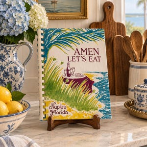 Peut inclure: Un livre de cuisine à reliure spirale intitulé Amen Let's Eat avec une illustration à l'aquarelle d'une table dressée au bord de l'eau, avec des palmes vertes, une nappe violette et le texte Naples Florida en police cursive décorative.