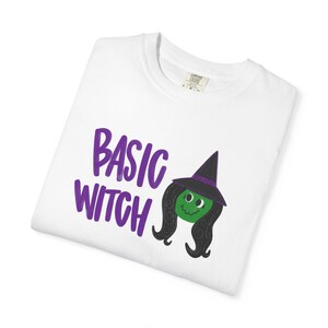 May include: A white t-shirt featuring the purple text "BASIC WITCH" next to a graphic of a green-faced witch with long black hair and a black pointed hat with a purple band. The shirt has a visible interior neck label with size L printed on it.