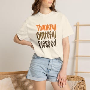 May include: A cream-coloured short-sleeve t-shirt featuring the stacked text Thankful in orange, Grateful in brown, and Blessed in dark brown. The shirt has a crew neckline and a relaxed fit, constructed from a solid fabric with no visible patterns.