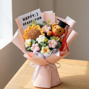 Puede incluir: Un ramo hecho a mano con un girasol, margaritas y flores rosas de ganchillo, con un oso de peluche con birrete. El ramo está envuelto en papel rosa y atado con una cinta blanca. La tarjeta dice "HAPPY :) GRADUATION".