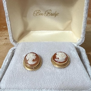 May include: A pair of oval-shaped cameo earrings featuring a white profile relief on a brown background, set in a gold-toned metal frame with a twisted rope border. The earrings are presented inside a light gray velvet-lined box with Ben Bridge text.