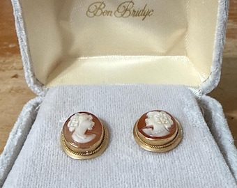 May include: A pair of oval-shaped cameo earrings featuring a white profile relief on a brown background, set in a gold-toned metal frame with a twisted rope border. The earrings are presented inside a light gray velvet-lined box with Ben Bridge text.