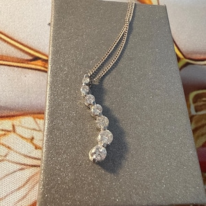 May include: A silver-toned necklace featuring a curved, vertical pendant set with six round, clear, faceted stones that graduate in size from top to bottom. The delicate chain is attached to the top of the stone-set pendant, creating a sleek, elegant look.