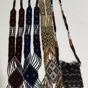 May include: A collection of beaded necklaces and a small beaded pouch. The necklaces feature intricate patterns in various colors, including burgundy, navy, gold, and black. Some have diamond-shaped pendants and fringe.