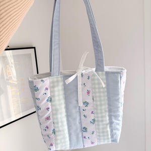 May include: A light blue quilted tote bag with floral and gingham patterned panels. The bag has matching light blue handles and a white ribbon bow. The bag is held up by a hand.