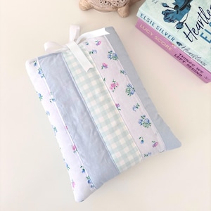 May include: A quilted tablet sleeve with a white ribbon tie. The sleeve has vertical stripes of floral fabric, light blue fabric, and a light blue gingham pattern. A ceramic turtle planter and two books are in the background.