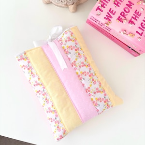 May include: A patchwork cushion with floral and polka dot fabric in pink, yellow, and white. A white ribbon is tied at the top. A pink book with the title "THINGS WE HIDE FROM THE LIGHT" is visible.