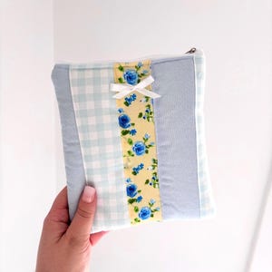 May include: A small, square, zippered pouch with a patchwork design. The pouch features light blue gingham, solid light blue, and a yellow floral stripe with blue roses. A white bow is attached to the floral stripe.