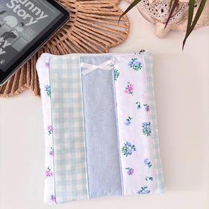 May include: A patchwork tablet sleeve with a charming design. It features sections of light blue gingham, solid light blue, and white floral fabric. A small white bow is attached. The text "Funny Story" is visible on a tablet.