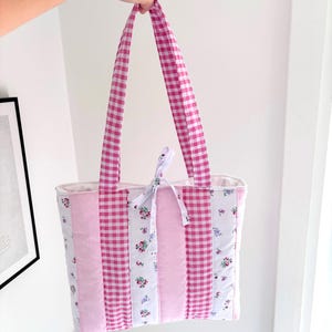 May include: A quilted tote bag featuring vertical panels of pink and white gingham, solid light pink fabric, and white fabric with a small floral print. The bag includes two matching pink gingham shoulder straps and a small tie closure at the top.