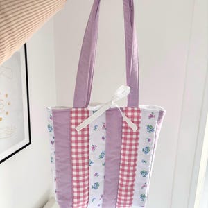 May include: A quilted tote bag with a patchwork design. The bag features vertical panels of lavender, pink gingham, and floral fabric. The handles and top trim are lavender. A white ribbon is tied at the top. The bag is held up by a person.