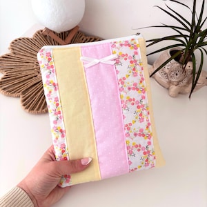 May include: A quilted pouch with a zipper closure. The design features vertical stripes of pale yellow, pink polka dots, and floral fabric with pink and yellow flowers. A small white bow is attached to the front. The pouch is held in a hand.