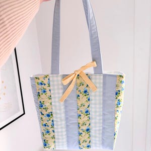 May include: A light blue tote bag with a floral and gingham pattern. The bag features a yellow bow and light blue handles. The floral pattern includes blue roses on a yellow background.