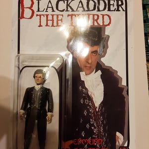 May include: A Blackadder the Third action figure in a clear plastic package. The packaging features a portrait of Edmund Blackadder and the show's logo. The figure is dressed in a black suit with a white shirt and vest.