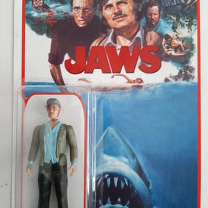 May include: A vintage 'Jaws' action figure of Quint, in a clear plastic package. The packaging features the film's poster art, including the title and a shark. The figure is wearing a cap and jacket.
