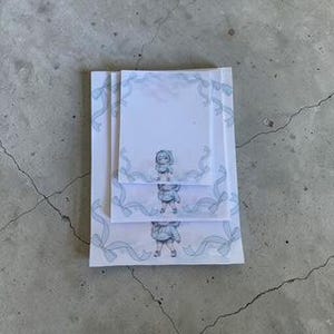 May include: Three white notepads of varying sizes, stacked on a gray surface. Each notepad features a light blue ribbon design around the edges and a small illustration of a character in the center.
