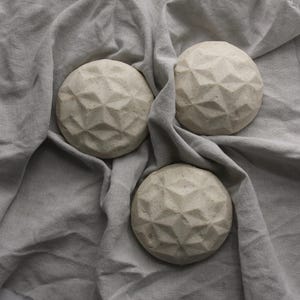 May include: Three circular, off-white objects with a raised, geometric, faceted star pattern on their top surfaces. Each object has a matte, textured finish and a dome-like shape, arranged in a cluster on a soft, light gray, wrinkled fabric.