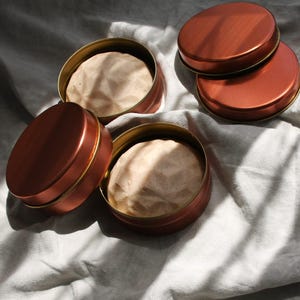 May include: Three round copper-colored metal tins, two of which are open to reveal beige, faceted, geometric-shaped solid bars inside. The third tin is closed with its matching copper-colored lid placed beside the open containers.