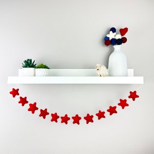 May include: A white shelf with a red felt star garland hanging below. The shelf is decorated with two small potted succulents and a small white stuffed sheep. A white vase with a bouquet of red, white, and blue felt balls sits on the shelf.