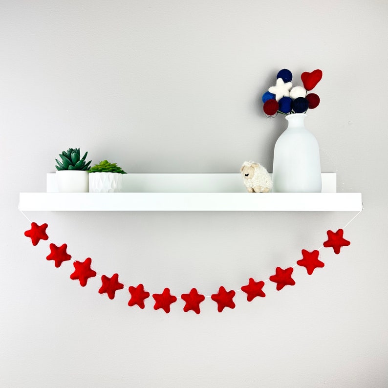 May include: A white shelf with a red felt star garland hanging below. The shelf is decorated with two small potted succulents and a small white stuffed sheep. A white vase with a bouquet of red, white, and blue felt balls sits on the shelf.