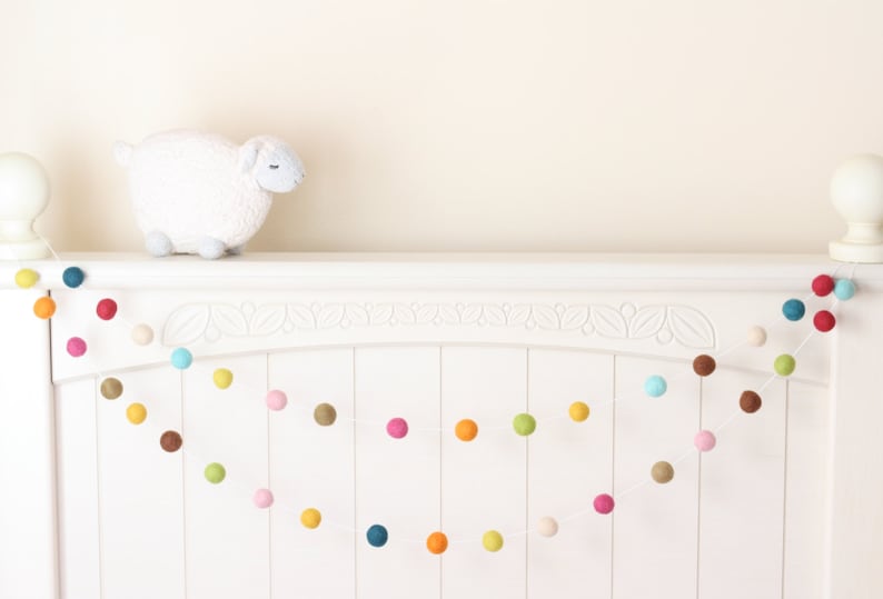 May include: A white headboard with a garland of colorful felt balls strung across it. The garland is made of various colors, including pink, blue, green, yellow, and orange.
