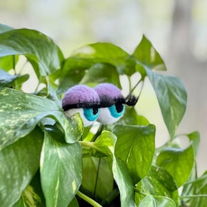 Peut inclure: Deux grands yeux en feutre avec des paupi&egrave;res violettes et des iris bleus qui d&eacute;passent de derri&egrave;re des feuilles vertes.