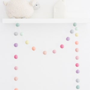 May include: A pastel colored felt ball garland hangs from a white shelf. The garland is made of soft, colorful felt balls in shades of pink, purple, green, and yellow. The garland is perfect for adding a touch of whimsy to any room.