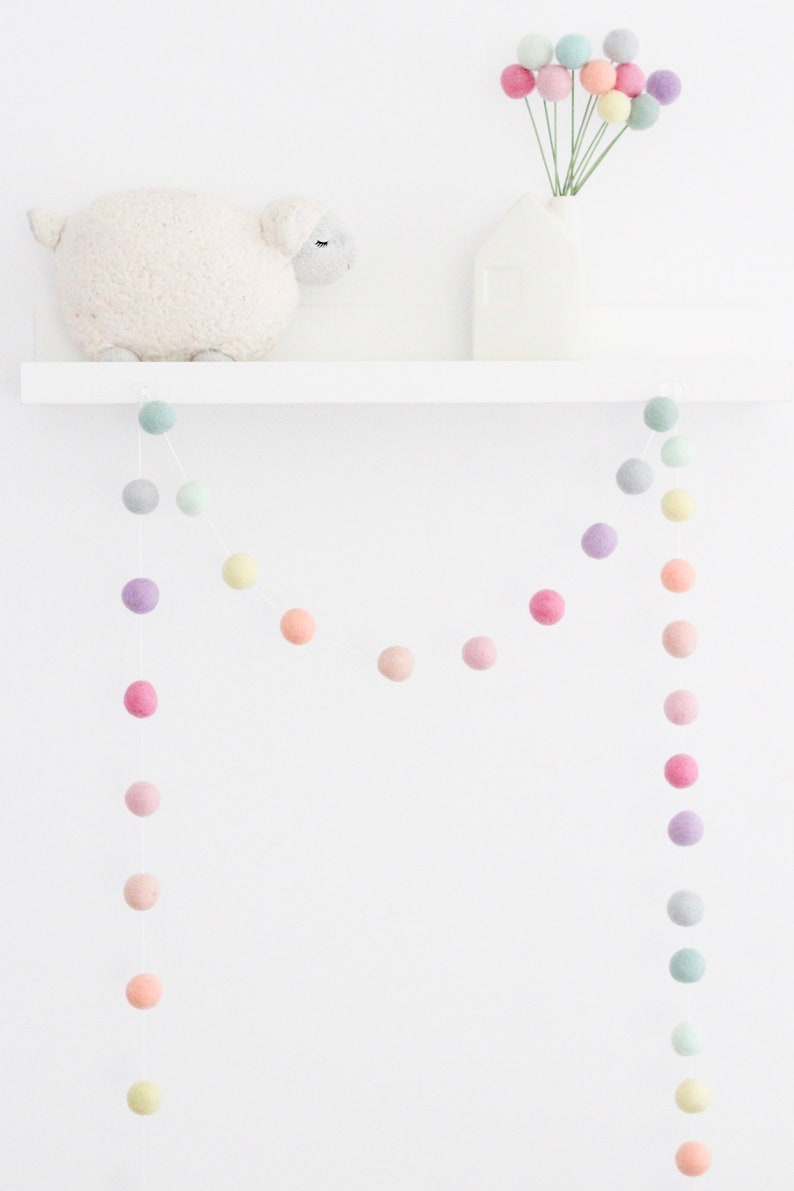 May include: A pastel colored felt ball garland hangs from a white shelf. The garland is made of soft, colorful felt balls in shades of pink, purple, green, and yellow. The garland is perfect for adding a touch of whimsy to any room.