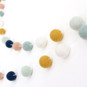 May include: A garland of felt balls in various colors, including blue, white, yellow, and pink. The balls are strung together with white thread and are arranged in a circular pattern.
