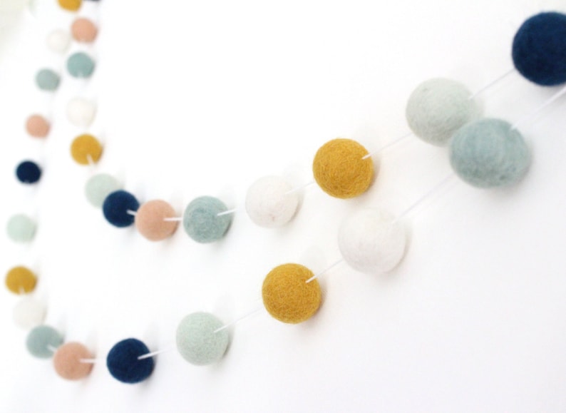 May include: A garland of felt balls in various colors, including blue, white, yellow, and pink. The balls are strung together with white thread and are arranged in a circular pattern.