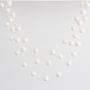 May include: A white felt ball garland with multiple strands of balls hanging from a white background.