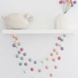 May include: A pastel colored felt ball garland hangs from a white shelf. The garland is made of soft, colorful felt balls strung together on a white string. The garland is perfect for adding a touch of whimsy to any room.