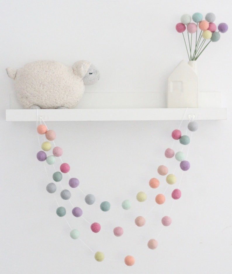 May include: A pastel colored felt ball garland hangs from a white shelf. The garland is made of soft, colorful felt balls strung together on a white string. The garland is perfect for adding a touch of whimsy to any room.