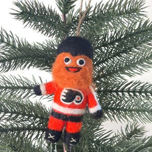 Op de afbeelding: Een naaldvilten ornament in de vorm van een ijshockeyspeler die een oranje, zwart en wit shirt draagt met een zwart logo op de borst. Het figuurtje heeft een pluizig oranje gezicht, een zwarte muts en zwarte schaatsen met witte veters.
