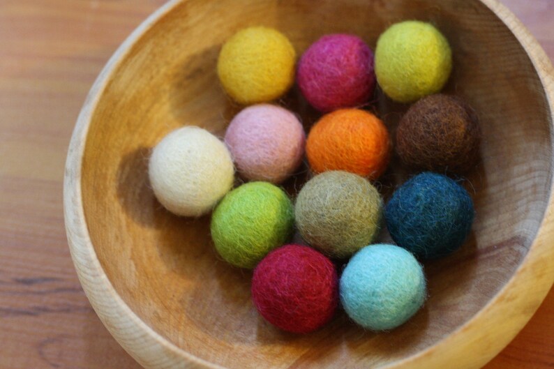 May include: A wooden bowl filled with colorful felt balls. The balls are in various shades of pink, green, blue, yellow, orange, brown, and beige.