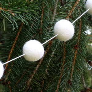 May include: A white felt ball garland strung on a white string, hanging on a green evergreen tree branch.