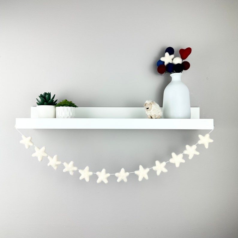 May include: A white shelf with a white felt star garland hanging from it. There are two small potted plants and a small white stuffed sheep on the shelf. A white vase with a bouquet of red, white, and blue felt balls is on the shelf.